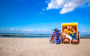 Paar entspannt in einem gelb-weißen Strandkorb mit zwei Fahrrädern daneben am Sandstrand in Wustrow unter blauem Himmel.