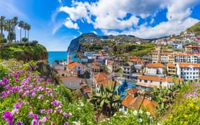 Bunte Häuser mit Terrakottadächern im Dorf Camara de Lobos an Madeiras Küste, umgeben von üppiger Vegetation und Bergen unter blauem Himmel.