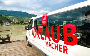 White Radweg-Reisen Urlaubmacher van with red lettering and bicycles inside, parked on wooden dock by river with hills and village in background.