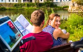 Two cyclists on a bench by the Danube River near Rechtenstein. One holds a map while the other smiles at the camera. Green vegetation and buildings visible.