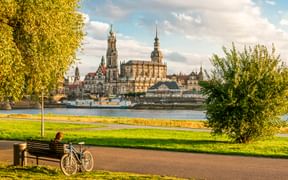 Person auf Bank mit Fahrrad auf Weg am Elbufer. Dresdner historische Skyline mit Domtürmen sichtbar über dem Fluss.