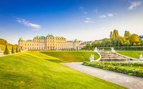 Schloss Belvedere in Wien mit symmetrischen Barockgärten, weißen Statuen, Brunnen und gepflegten Rasenflächen unter blauem Himmel.