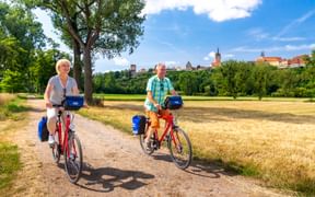 Zwei Radfahrer auf roten Tourenrädern mit blauen Taschen auf einem Weg bei Bad Wimpfen. Historische Stadtsilhouette mit Kirchtürmen im Hintergrund.