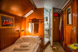 Kompakte Kabine auf MS L'Estello mit holzgetäfelten Wänden, Einzelbett mit weißer Bettwäsche, Wandlampe und eigenem Bad durch offene Tür sichtbar.