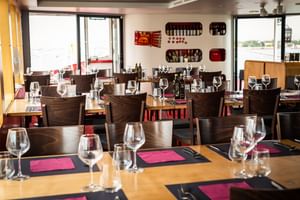 Bright restaurant interior with wooden tables set with wine glasses and pink napkins. Wall shelves display wine bottles. Large windows overlook water.