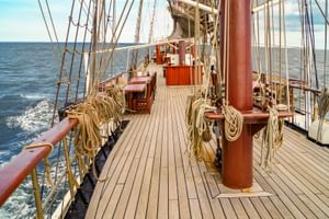 Holzdeck des Großseglers Atlantis mit Mast, Takelage und aufgerollten Seilen. Das Schiff segelt auf blauem Meer unter teils bewölktem Himmel.