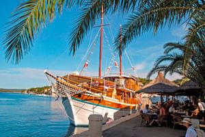 Traditionelles Holzsegelschiff MS Orkan mit weiß-blauem Rumpf an einer Uferpromenade vertäut. Palmen rahmen die Szene, eine Café-Terrasse ist sichtbar.