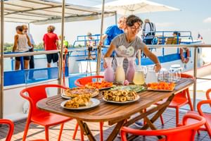 Crew-Mitglied serviert Essen und Getränke auf dem Sonnendeck der MS Caprice. Tisch mit Gebäck, Obst und Getränken. Passagiere entspannen auf blau-weißem Bootsdeck.