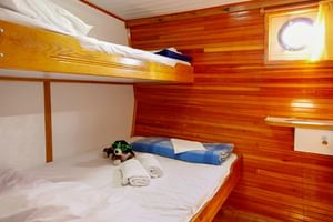 Holzkabine auf der MS Mirabela mit Etagenbetten, weißer Bettwäsche, blauen Handtüchern und rundem Bullauge mit gelben Vorhängen.