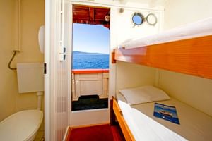 Kompakte Kabine auf der MS Dalmatino mit Holz-Etagenbetten, weißer Bettwäsche und eigenem Bad. Offene Tür zeigt Deck mit Meerblick und Bullaugen.