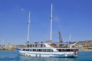 Weiße Motoryacht MS Aria mit blauem Zierstreifen und zwei Masten im Hafen. Weiße Fender entlang des Rumpfes, Küstenstadt im Hintergrund.