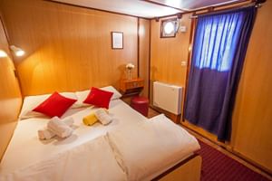 Gemütliche Kabine auf der MS Azimut mit Doppelbett, roten Kissen, holzgetäfelten Wänden, Bullauge mit blauen Vorhängen und weißer Heizung.