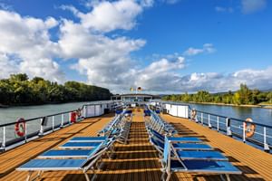 Sonnendeck der MS Olympia mit Reihen blauer Liegestühle auf Holzboden, weißen Geländern und Flusslandschaft im Hintergrund.