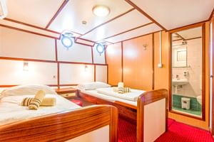 Doppelkabine auf dem Schiff Amore mit zwei Einzelbetten, Holzmöbeln, Bullaugen, rotem Teppich und eigenem Bad mit Waschbecken.