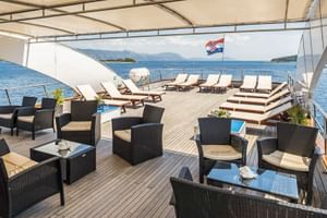 Geräumiges Sonnendeck der MS Olmissum mit Holzboden, Sonnenliegen, Korbmöbeln und kroatischer Flagge. Küstengebirge im Hintergrund sichtbar.