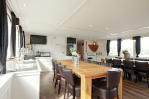 Geräumiger Salon an Bord der MS Magnifique II mit großem Holzesstisch, braunen Lederstühlen und weißem Küchenbereich. Große Fenster mit Vorhängen an beiden Seiten.