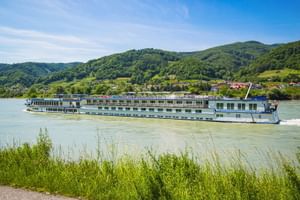 Weißes Flusskreuzfahrtschiff MS Swiss Crown auf der Donau mit grünen Hügeln, einem Dorf und blauem Himmel im Hintergrund.