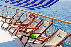 Reihe von Liegestühlen mit Mirabela-Logo auf dem Sonnendeck unter blau-weiß gestreifter Markise. Chromgeländer und ruhiges blaues Wasser im Hintergrund.
