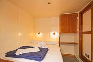 Kompakte Kabine auf der MS Sveti Vid mit weißem Doppelbett, blauen Handtüchern, Wandleuchten, Holzschrank und Tür mit Milchglasscheiben.