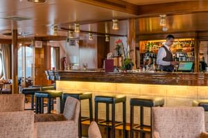 MS Olympia Schiffsbar mit beleuchtetem Holztresen, Barhockern, beigen Sesseln und Barkeeper. Bullaugen und warmes Licht schaffen gemütliche Atmosphäre.