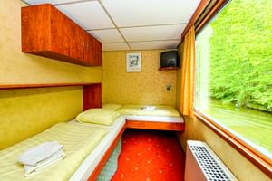 Kompakte Kabine auf der MS Fluvius mit zwei Betten, Holzschränken, TV und großem Fenster mit Blick auf grüne Uferlandschaft. Roter Teppich.