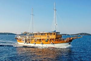MS Aneta, ein hölzernes Segelschiff mit zwei Masten und weiß-braunem Rumpf, fährt auf ruhigem blauen Wasser mit grünen Inseln im Hintergrund.