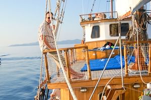 Holzdeck der MS Panagiota mit Geländer und Takelage. Eine Person steht an Deck, ruhige See und ferne Berge im Hintergrund sichtbar.