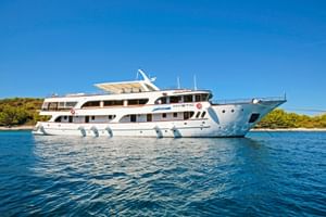 Weiße Motoryacht MY Mystic ankert in ruhigem blauen Wasser mit grüner Küstenlinie im Hintergrund unter klarem Himmel.