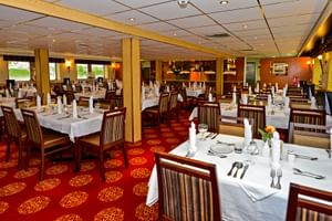 Elegantes Restaurant an Bord der MS Normandie mit weiß gedeckten Tischen, Holzstühlen, rot gemustertem Teppich und warmer Beleuchtung.