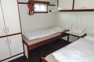 Zweibettkabine auf der MS Lena Maria mit zwei Einzelbetten, weißen Wänden, dunklem Teppich, Einbauschrank und kleinem Fenster über den Betten.