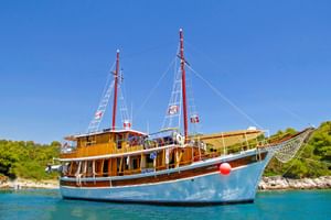 MS Orkan, ein Holzkreuzfahrtschiff mit blauem Rumpf und zwei roten Masten, vor Anker in türkisfarbenem Wasser nahe bewaldeter Küste unter blauem Himmel.