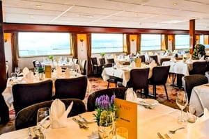 Eleganter Speisesaal an Bord der MS Rigoletto mit weiß gedeckten Tischen, schwarzen Stühlen und großen Fenstern mit Flussblick.