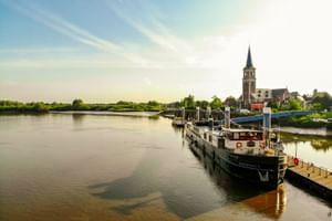 Flusskreuzfahrtschiff MS Magnifique II an einem Holzsteg vertäut. Eine Kirche mit hohem Turm steht am gegenüberliegenden Flussufer unter blauem Himmel.