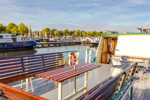 Sonniges Deck der MS Flora mit Holzbänken, weißem Geländer und Rettungsring. Marina mit Booten und Bäumen im Hintergrund unter blauem Himmel.