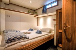 Gemütliche Doppelkabine auf der MS Flora mit gemachtem Bett, gestreifter Decke, Handtüchern, Holzschrank und Fenster mit Vorhang.