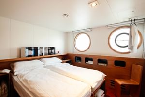Bright double cabin on MS Ave Maria with white bedding, wooden paneling, two round portholes, and a small seating area.