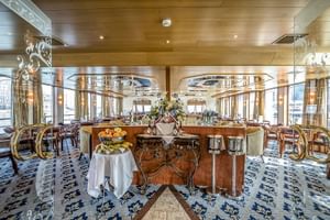 Elegante Panoramabar auf der MS Swiss Crown mit Holzdecke, großen Fenstern, gemustertem Teppich und Buffettisch mit frischem Obst und Blumen.