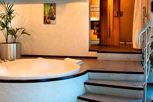 Wellnessbereich auf der MS SE-Manon mit rundem Whirlpool, Heimtrainer, grünen Pflanzen und Holzsauna im Hintergrund.