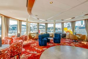 Geräumige Lounge auf der MS Lisabelle mit rot gemustertem Teppich, blauen und orangen Sesseln und großen Fenstern mit Hafenblick.