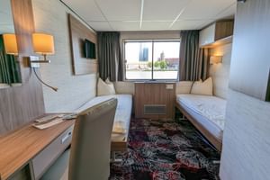 Kompakte Doppelkabine auf der MS Normandie mit zwei Einzelbetten, Holzverkleidung, Schreibtisch, Stuhl, Wandfernseher und Fenster mit Stadtblick.