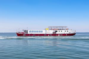 MS Ave Maria river cruise ship with red and white hull sailing on calm blue water under clear sky. The vessel features multiple decks and portholes.