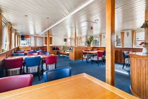Geräumiges Salon-Restaurant an Bord der Floor van Aemstel mit Holzvertäfelung, blauem Teppich, roten und blauen Stühlen unter weißer Decke.
