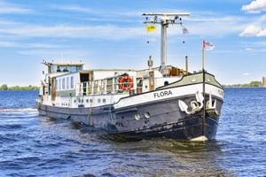 MS Flora, ein weiß-schwarzes Fahrgastschiff mit blauen Fenstern, fährt auf ruhigem Wasser unter blauem Himmel mit weißen Wolken.