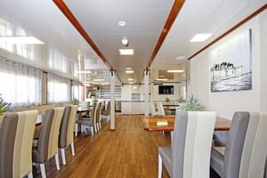 Heller Speisesaal auf der MS Aria mit Holzboden, mehreren Tischen mit beigen und grauen Stühlen, weißen Säulen und Pantryküche im Hintergrund.