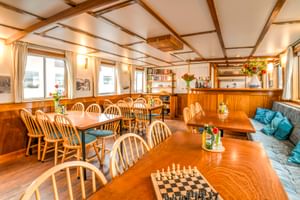 Heller Salon an Bord der MS Flora mit Holzdeckenbalken, Esstischen mit Windsor-Stühlen, Schachspiel, eingebauter Bar und blauer Polsterung.