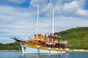 MS Planka, ein hölzernes Zweimaster-Segelschiff mit weißem Rumpf und gelb-braunem Aufbau, vor Anker in ruhigem blauen Wasser mit grünen Hügeln.