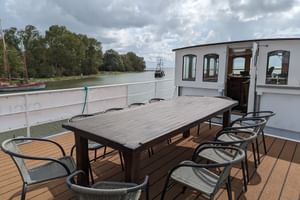 Esstisch mit Stühlen auf dem Sonnendeck des Schiffes Floor van Aemstel, mit Flussblick und Steuerhaus im Hintergrund unter bewölktem Himmel.