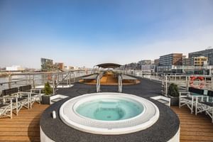 Sonnendeck der MS Swiss Crown mit rundem Whirlpool, Liegestühlen und Holzdeck. Städtische Uferpromenade mit modernen Gebäuden im Hintergrund.