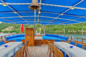 Sonnendeck der MS Thalassa mit Holzbänken, blauen Kissen und blauer Markise. Küstenlandschaft mit grünen Hügeln im Hintergrund.