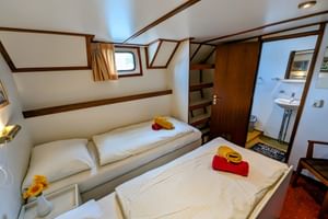 Zweibettkabine auf Floor van Aemstel mit zwei Einzelbetten, weißer Bettwäsche, roten Handtüchern, gelben Hüten, Holzverkleidung, Bullauge und eigenem Bad.